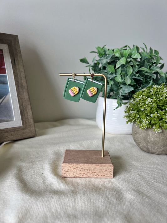 Green earrings on a gold stand with a wooden base, placed on a surface with plants in the background.